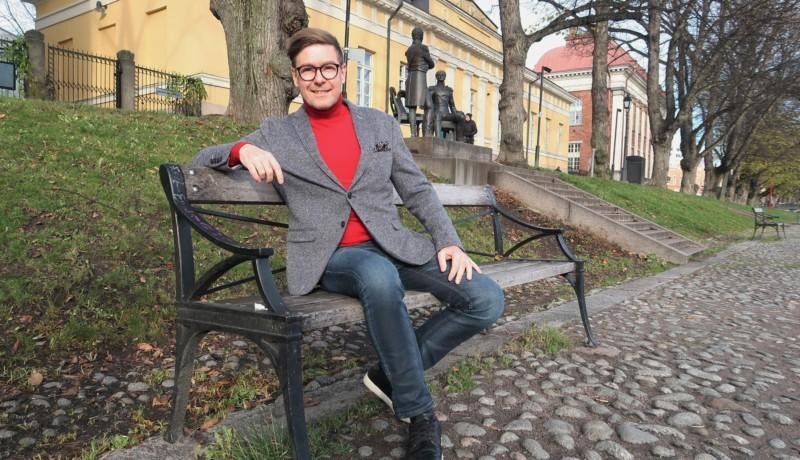 Bild av en man i bleiser och polotröja som sitter på en bänk vid åstranden i Åbo.