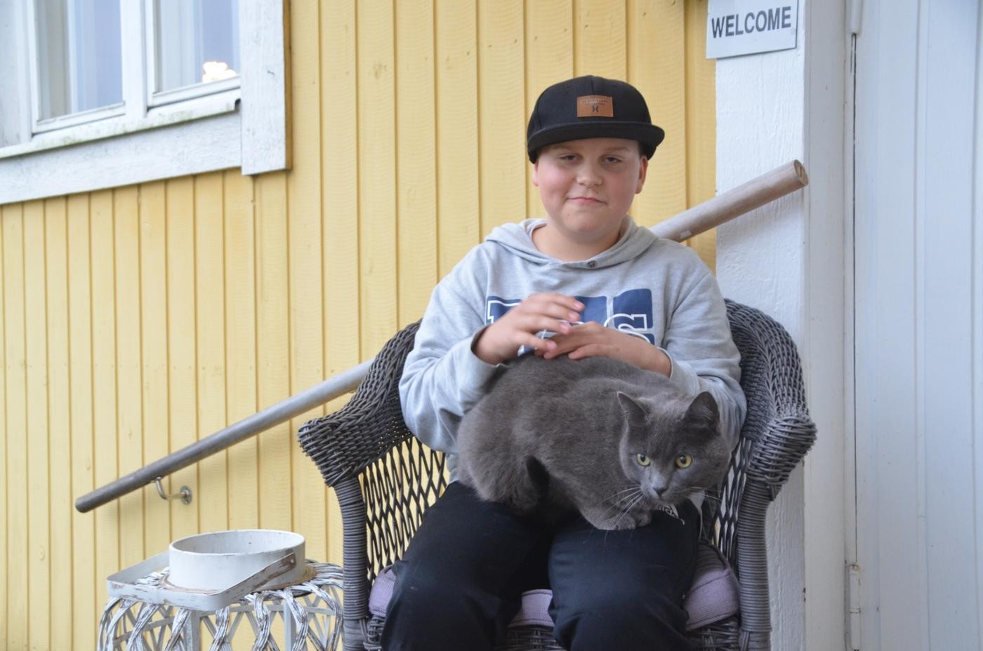 pojke sitter utomhus i korgstol och har en katt i famnen