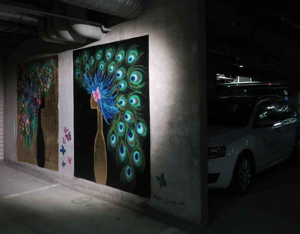 Bild av konst som föreställer ett huvud med en krona av påfågelsfjädrar på en vägg i en parkeringshall. Bakom konstverket skymtar en parkerad bil.