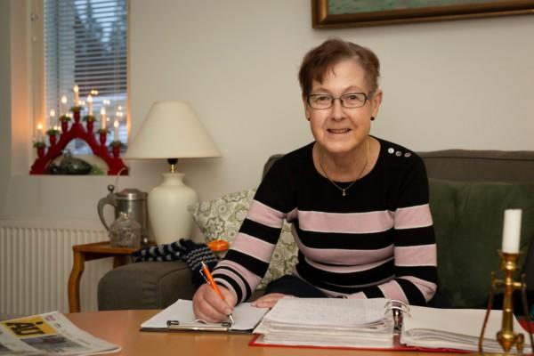 äldre kvinna sitter i soffa med papper och penna framför sig på soffbordet