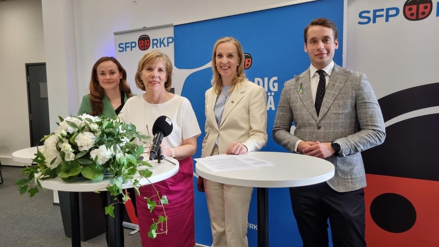 tre kvinnliga politiker och en manlig på en presskonferens, stor vit blomuppsättning framför dem