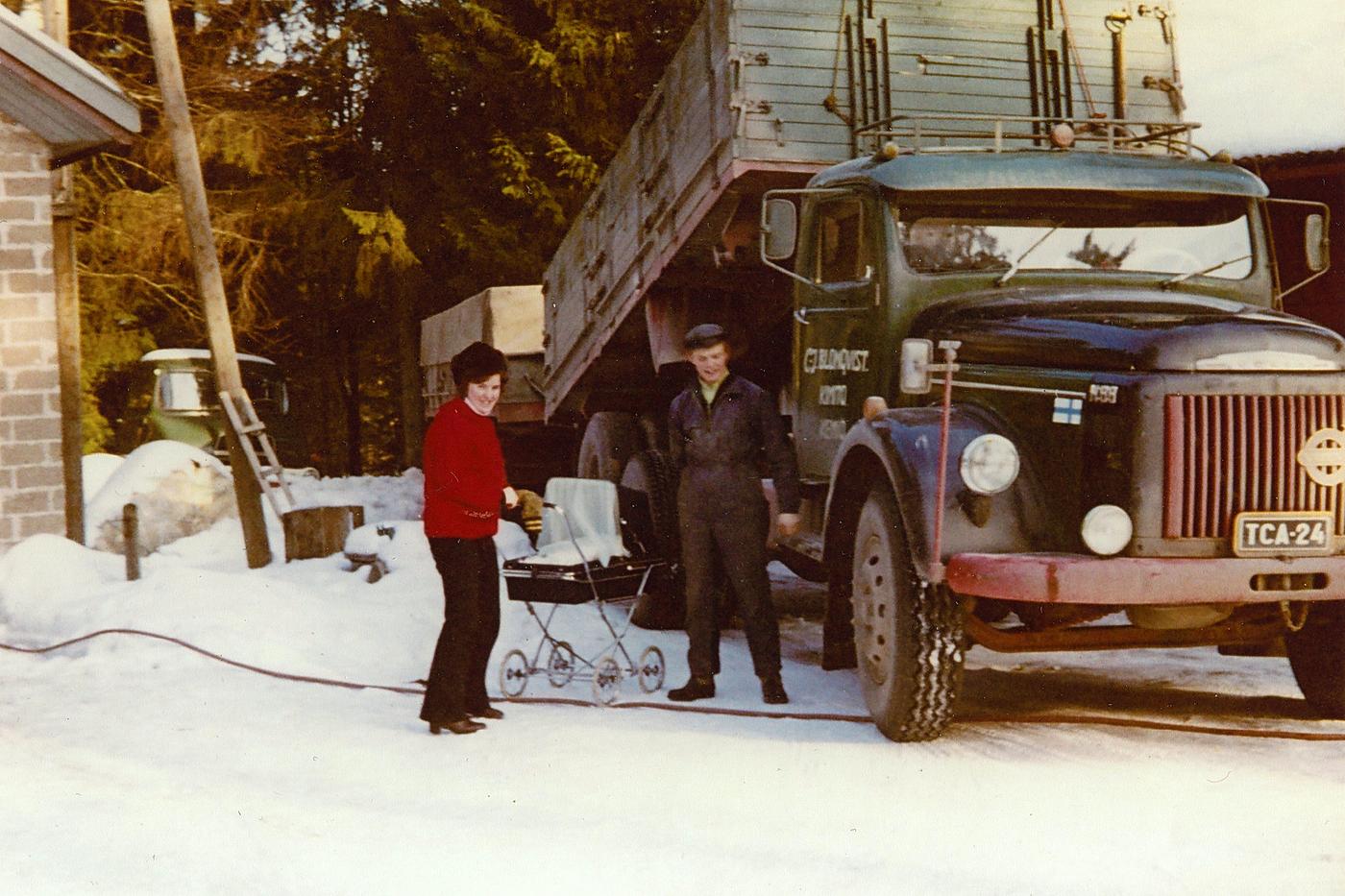 Ett gammalt fotografi föreställande en kvinna och en man med baby i en vagn och en lastbil i bakgrunden.