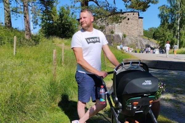 En man iklädd sommarkläder står utomhus med en barnvagn.