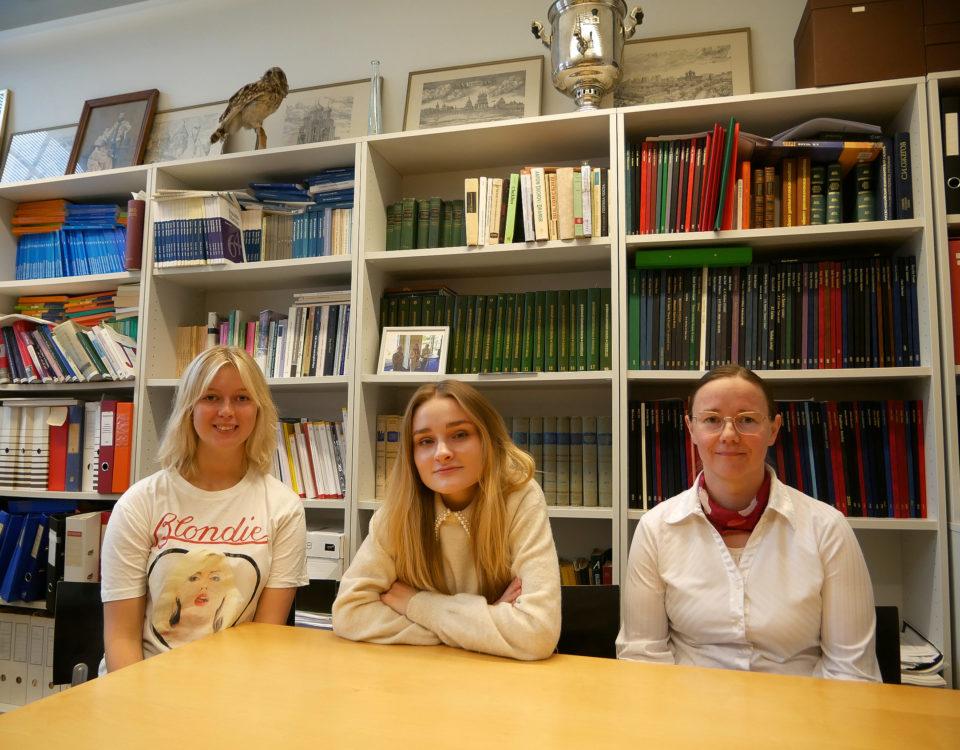 Tre studerande sitter vid bord framför bokhylla.