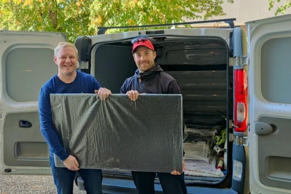 Två leende män håller upp en inpaketerad tv framför en paketbil.