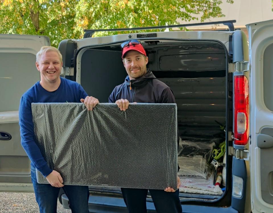 Två leende män håller upp en inpaketerad tv framför en paketbil.