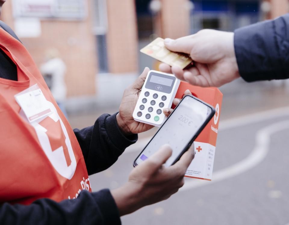 Person som håller i en insamlingsbössa och betalterminal, en annan persons hand sträcker sig från höger sida med ett bankkort i handen.