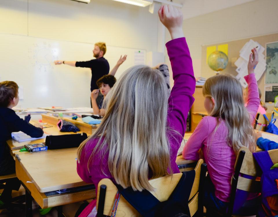 Flicka räcker upp handen och en lärare står vid tavlan.