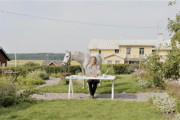 Kvinna sitter vid skrivbord utomhus, bakom står en vit häst.