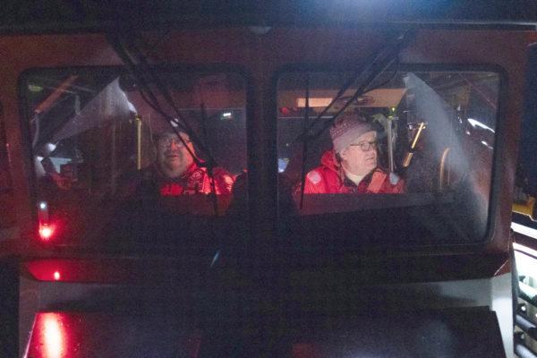 En sjöräddningsbåts förarhytt i mörker. Två män syns genom rutan.