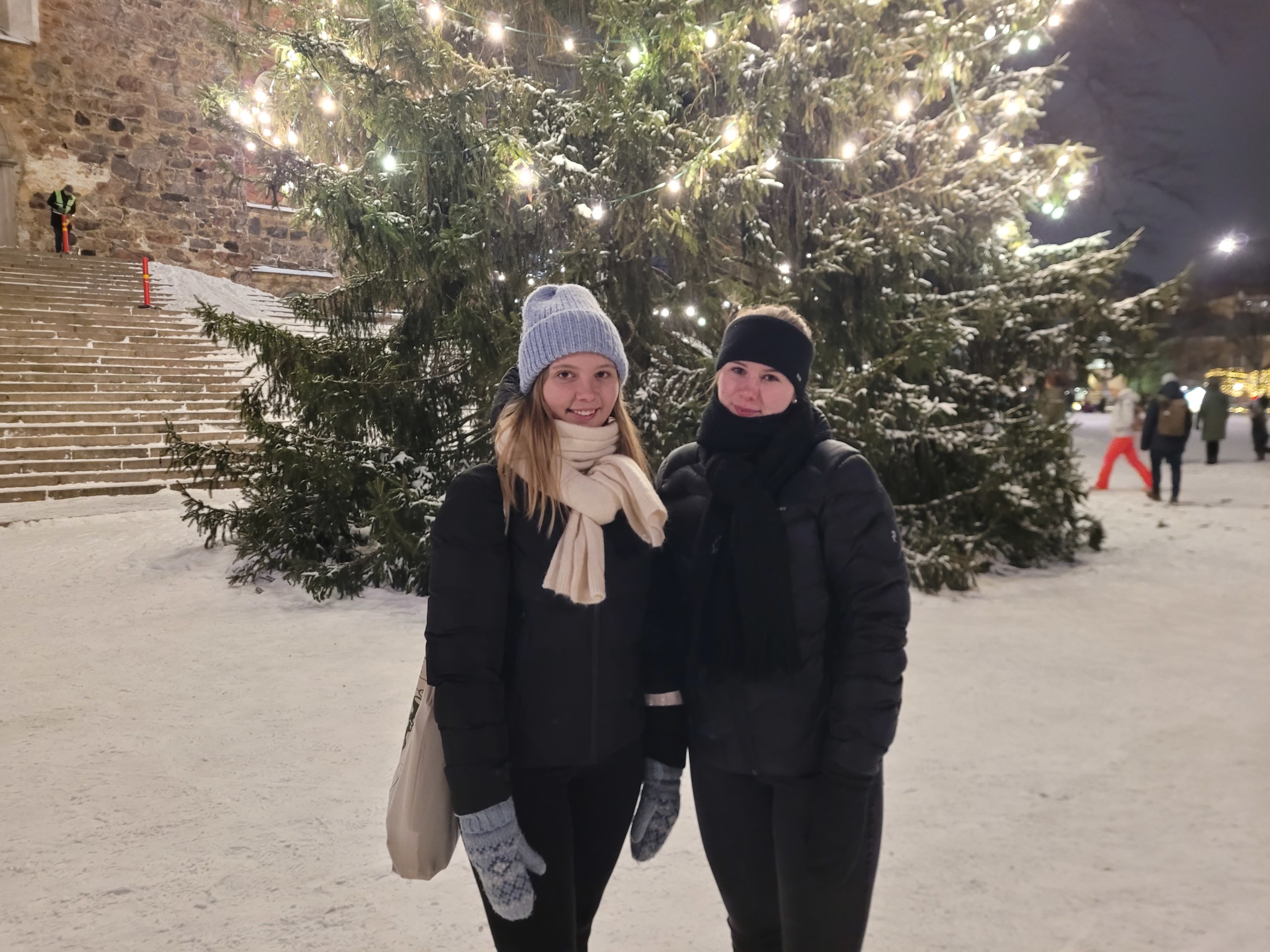 Två kvinnor står i snön framför en stor julgran.