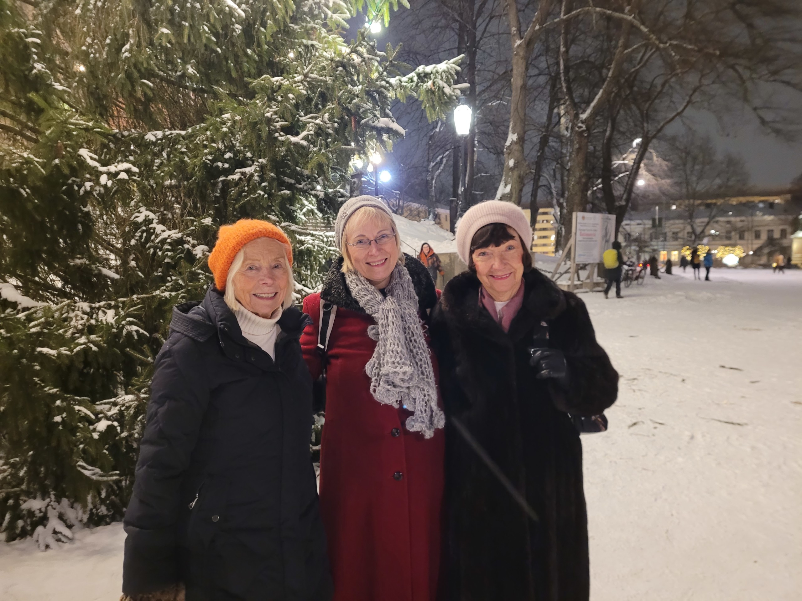 Tre damer står framför en julgran och ler mot kameran.