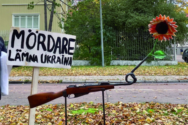 Ett gevär vars pipa har en knut på sig och en röd blomma stickande upp. Bredvid en skylt med texten "Mördare lämna Ukraina".