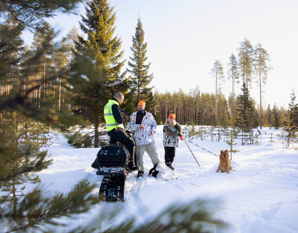 En inspektör iklädd reflexväst inspekterar en jägare. En flicka och en hund står bredvid dem i den snötäckta skogsgläntan.