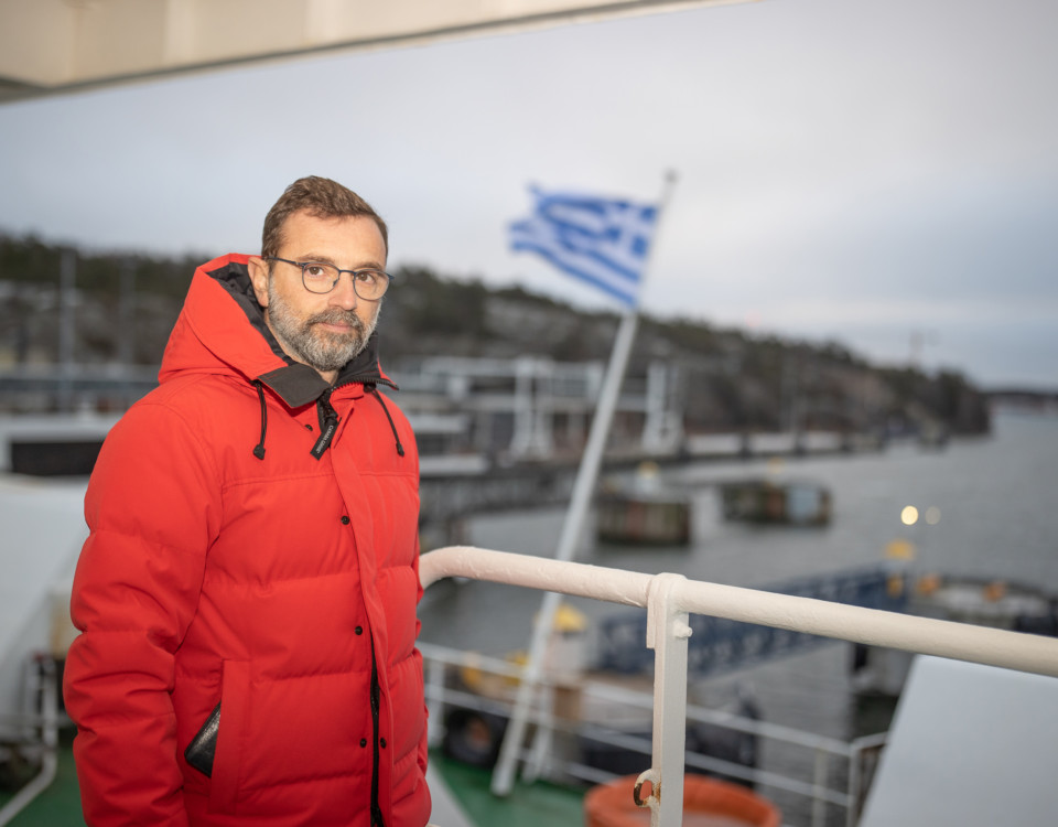 En man i röd täckjacka ombord på ett fartyg, i bakgrunden vajar Greklands flagga.