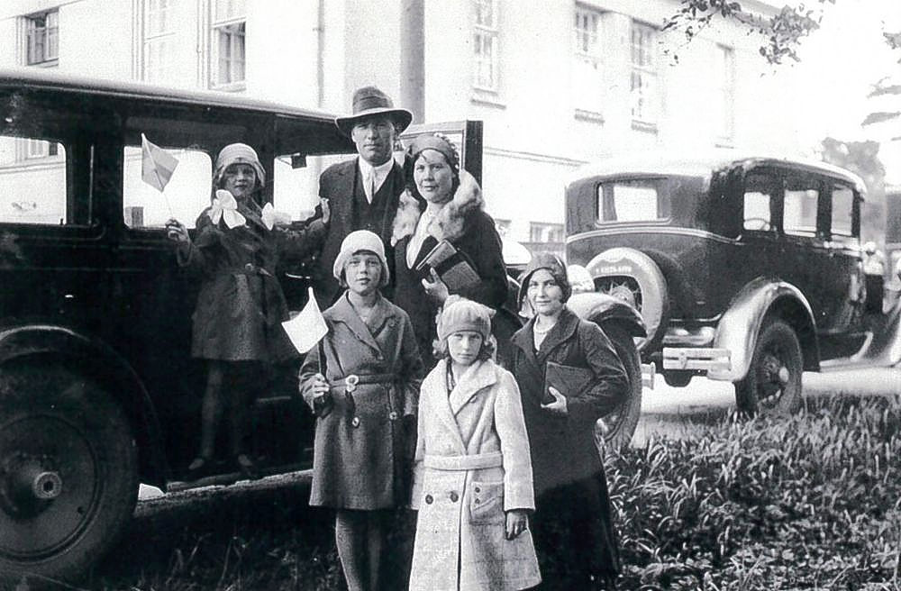 Ett svartvbitt fotografi föreställande en bil och en familj. 