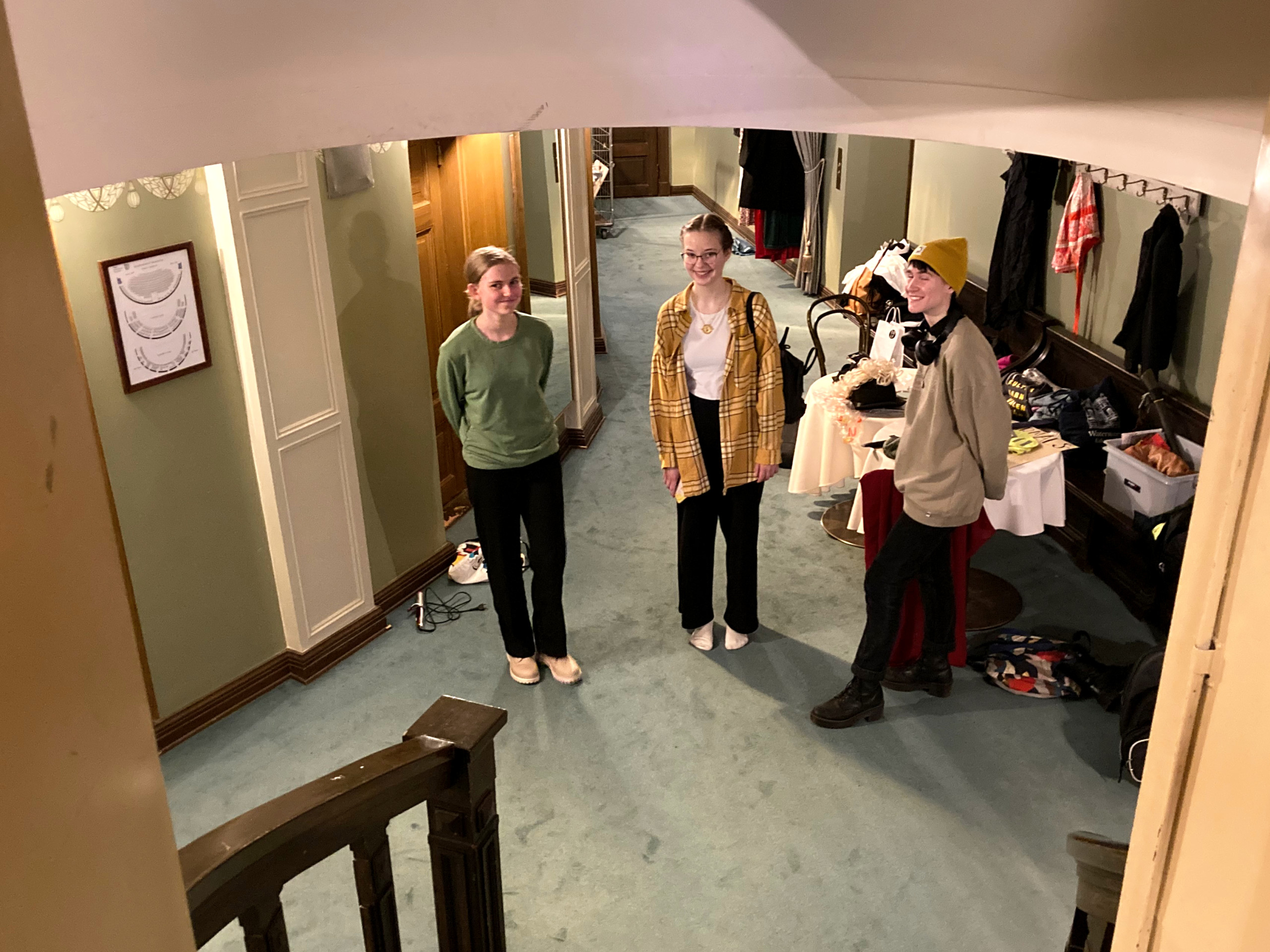 Tre unga personer i en teaterkorridor, bakom dem syns högar av kläder och väskor.