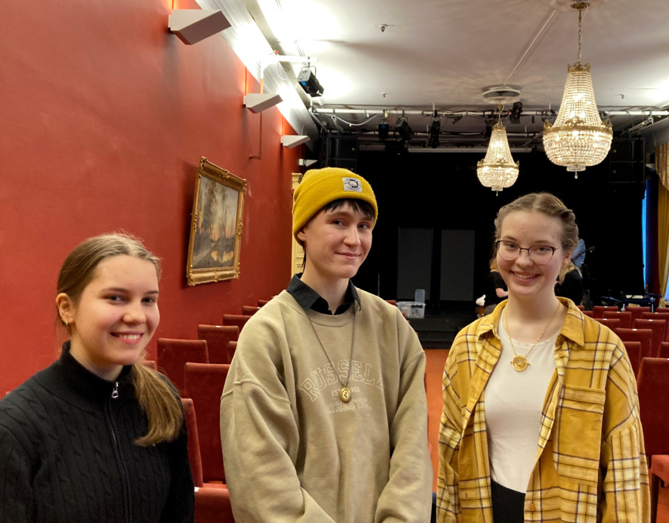 Tre unga personer i en teaterlokal med röda väggar och kristallkronor.