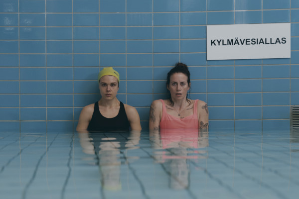 Två kvinnor i baddräkter sitter i en vattenbassäng med blå kakel. På väggen en skylt där det står "kylmävesiallas".