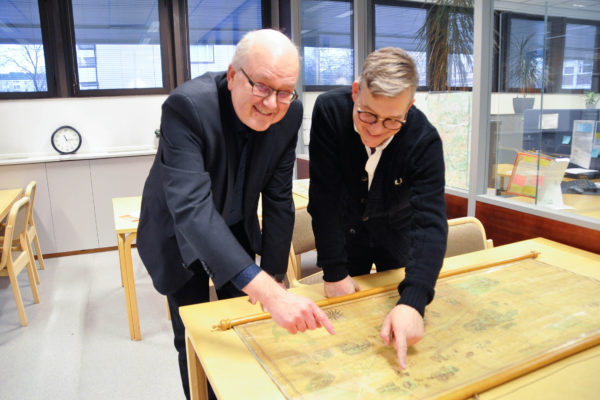 två män står vid ett bord med gulnat arkivmaterial och pekar på det