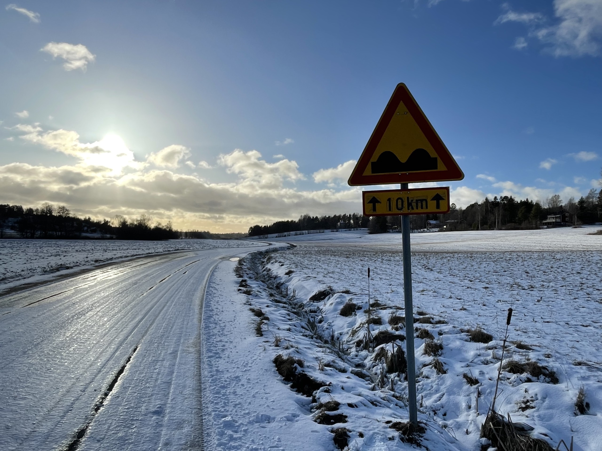 En istäckt väg, kantad av åkrar med ett varningsmärke om gropig väg. 