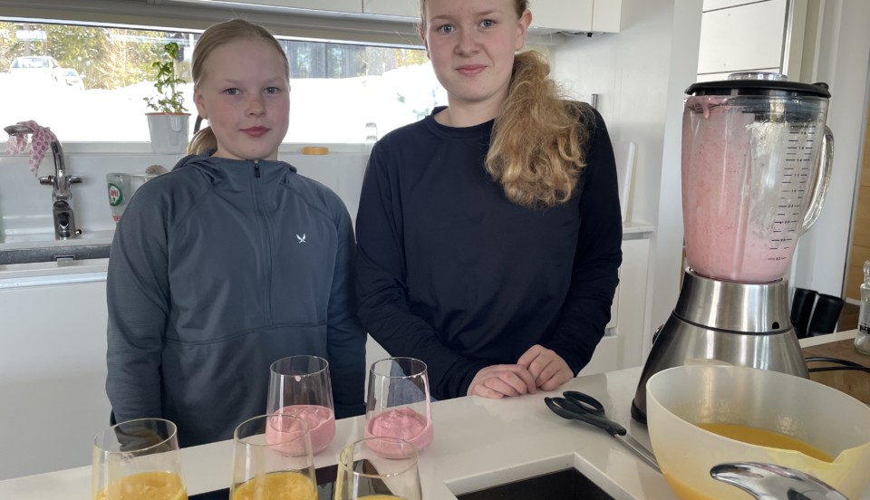 två flickor i ett kök. Flera glas med smoothie på bordet och en mixer.