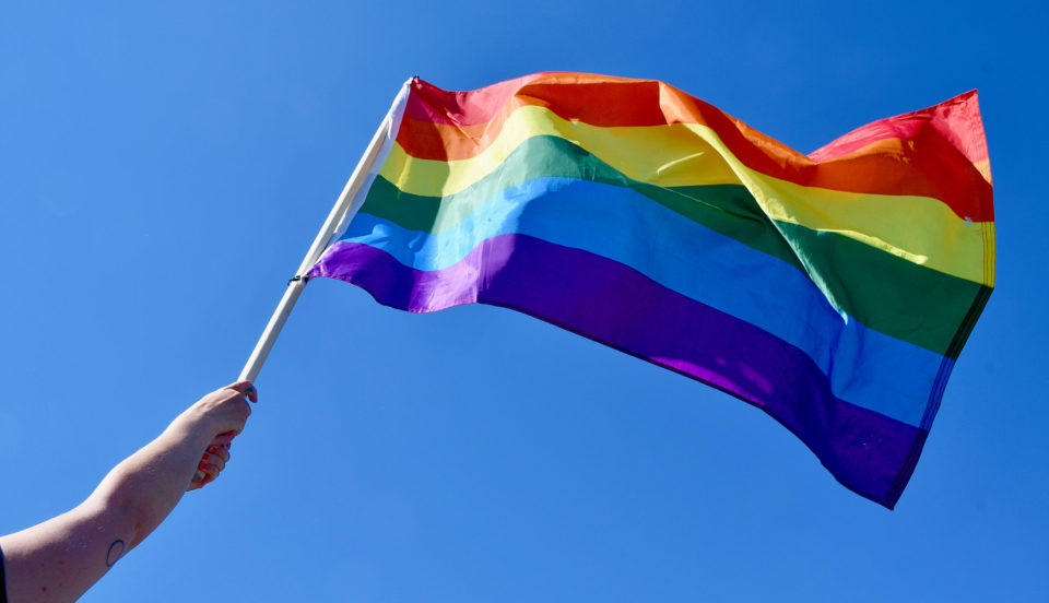En hand som håller upp en regnbågsflagga mot blå himmel