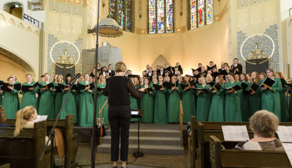 En kör bestående av både kvinnor och män framme i koret i en kyrka. En svartklädd dirigent framför dem.