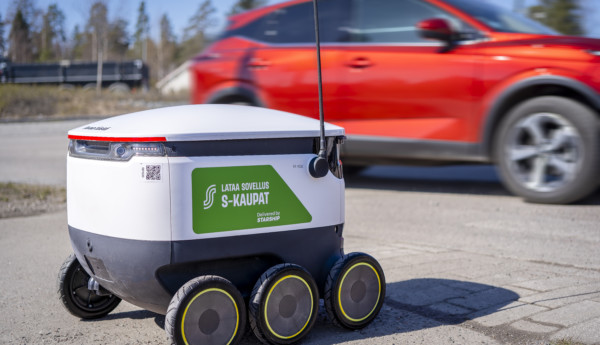 En lådformad robot på sex hjul, i bakgrunden kör en röd bil förbi