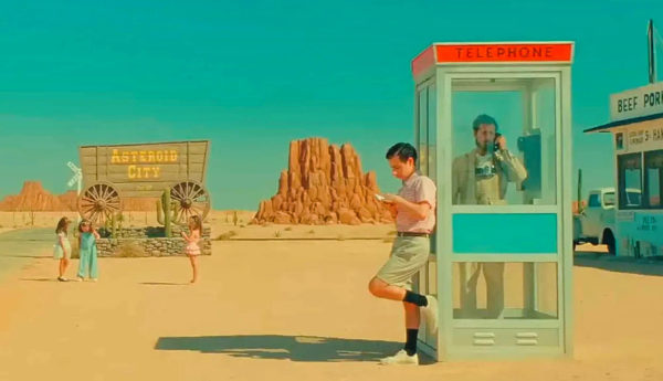 Ökenlandskap i 50-tals pastellfärger. En man står i en telefonkiosk, i bakgrunden en skylt i formen av en kärra med texten "Asterioid City".