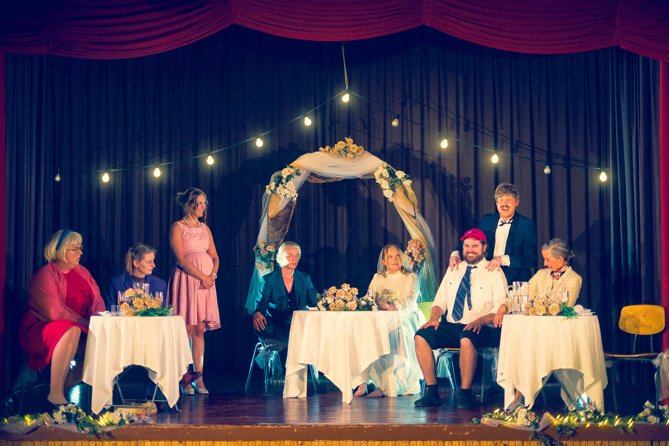 Skådespelare på scen i en bröllopsfest. Bord är dukade och brudparet sitter i mitten medan deras familjer är på varsin sida om dem vid egna bord.