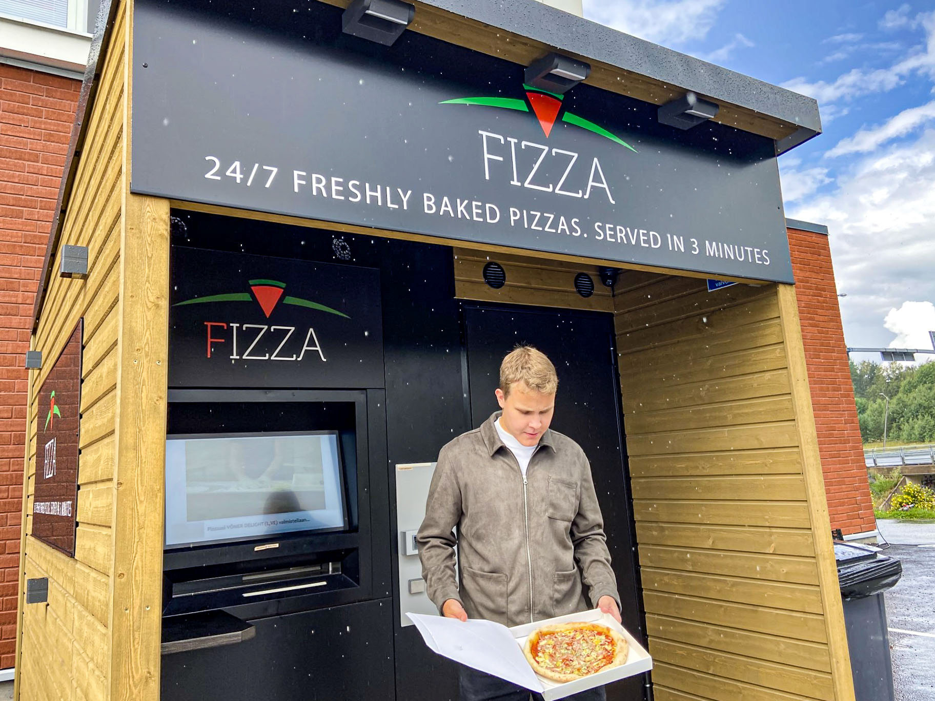 En man står framför pizzaautomaten. Han håller i en pizza som han ser på.