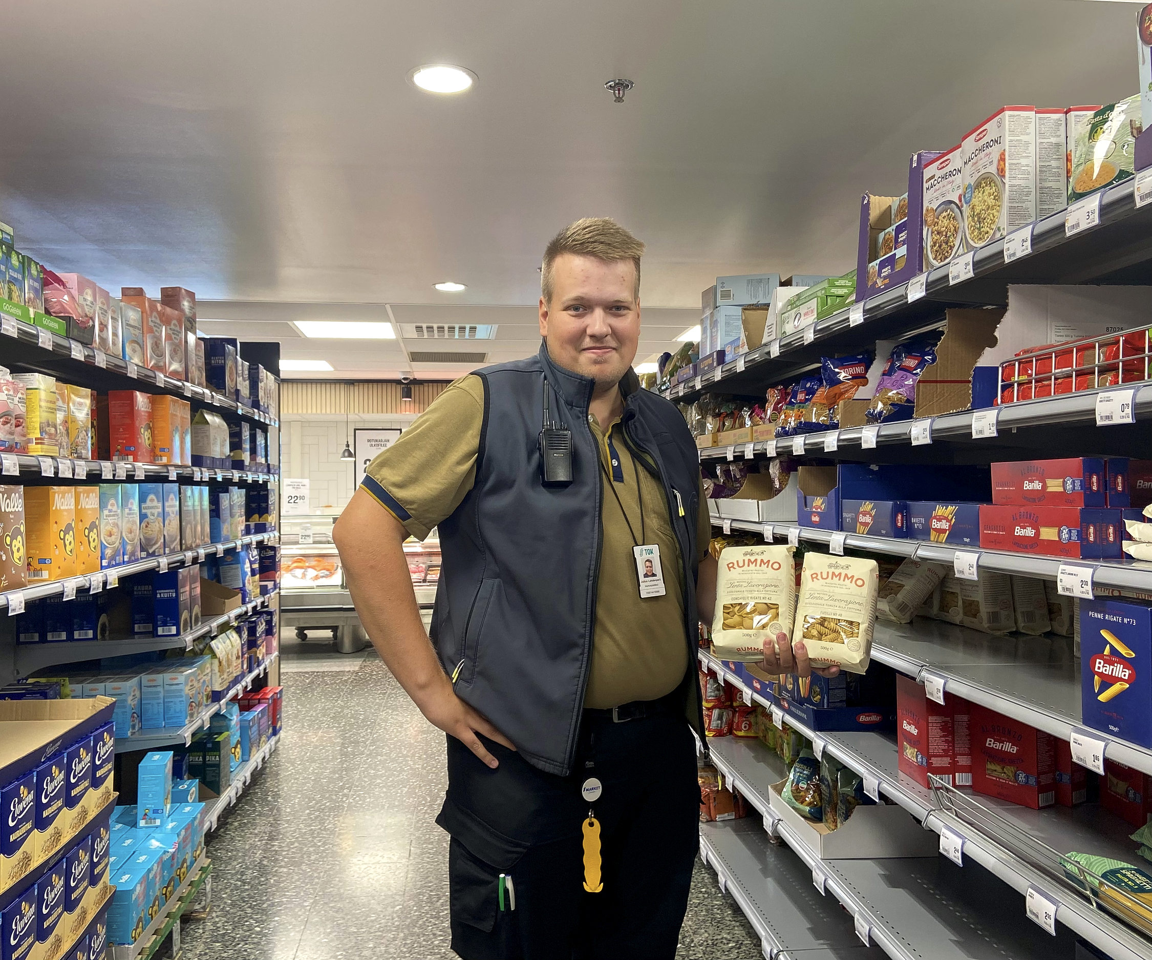 En man i arbetskläder och pastapaket i handen står mellan två butikshyllor.