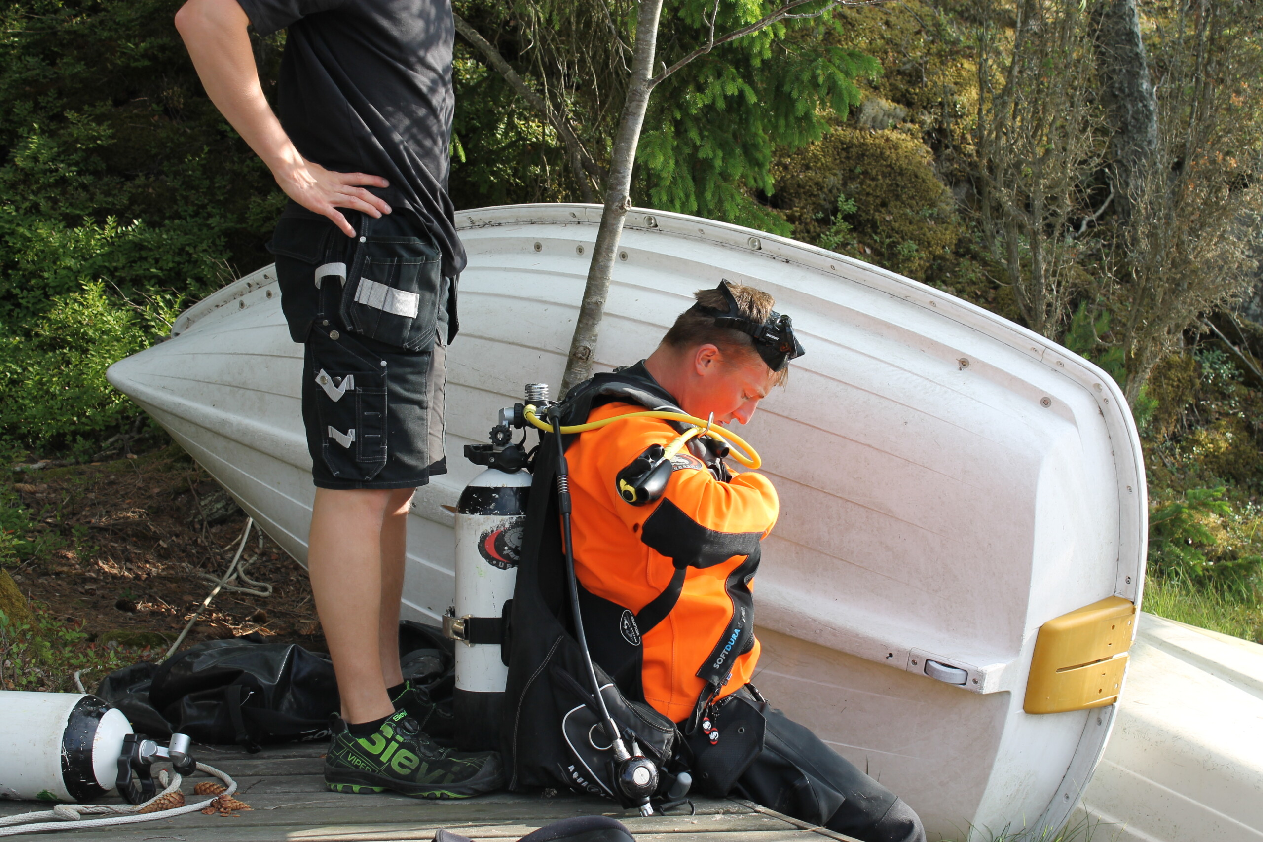 Ung man sitter på en brygga och sätter på sig sin dykarutrustning, en person står bakom honom och hjälper till att få tanken fast på ryggen.