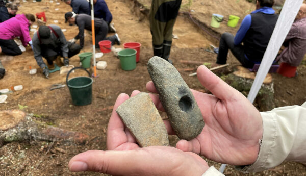 Två arkeologiska fyns (stenyxor) i ett handpar. I bakgrunden en utgrävningsplats.