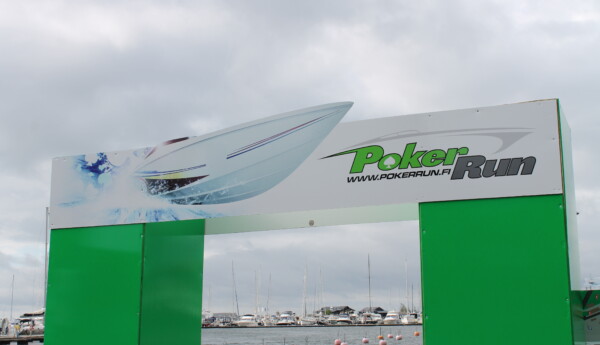 Närbild på en skylt i Hangö hamn med texten "Poker run" i bakgrunden syns vatten och båtar.
