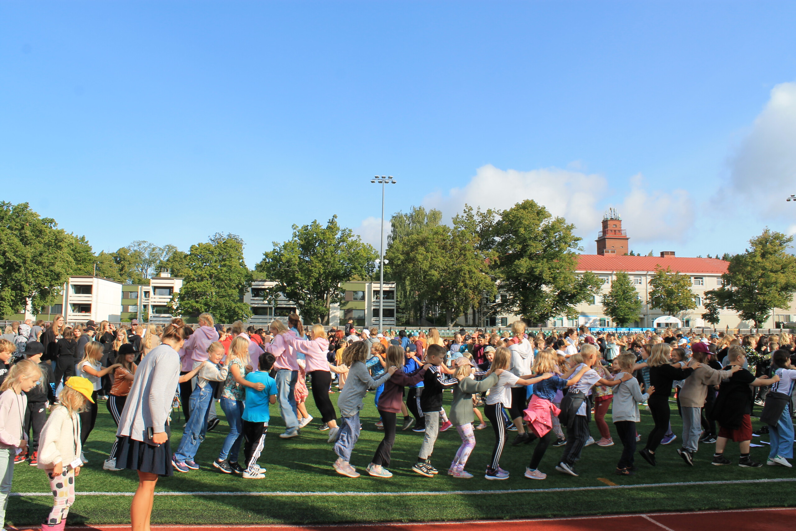 Elever och lärare är utomhus på en gräsplan och dansar letkajenkka. Eleverna är från skolorna Ekenäs högstadium, Seminarieskolan och Hakarinne i Ekenäs.