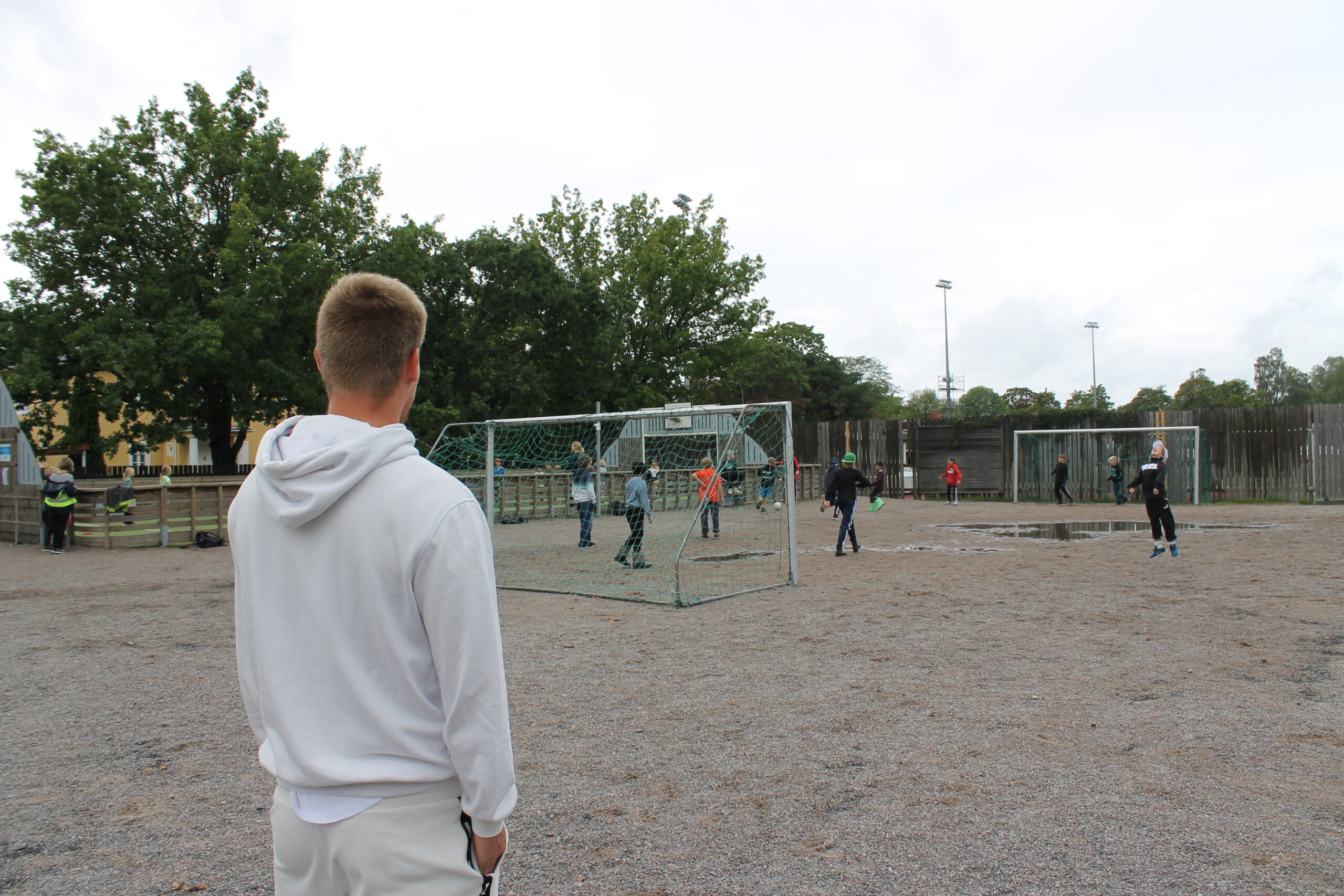 En ung man fotad med ryggen mot kameran, han står och ser på Seminarieskolans fotbollsplan på skolgården där några elever spelar fotboll.