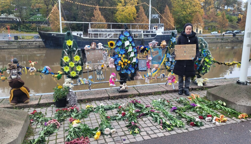 En kvinna står vid åstranden och håller i ett plakat med texten "Sluta döda" på engelska, svenska, finska och ryska. Runtomkring finns blommor, Ukrainaflaggor och andra föremål som visar stöd för Ukraina.