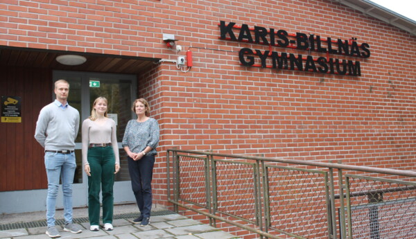 En man, en ung kvinna och en kvinna står bredvid varandra utanför en röd tegelbyggnad med skylten "Karis-Billnäs Gymnasium". De arbetar alla på skolans skoltidning.