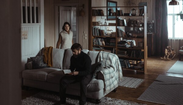 en man sitter i en soffa med en bok i handen och en kvinna står bakom soffan