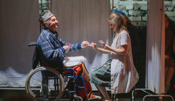 Teaterscen med man i rullstol och sköterska som spalr kort.