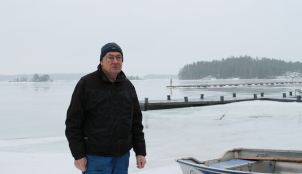En äldre man står utomhus vid hamnen Baggö i Ekenäs skärgård. Det är snö på marken och i bakgrunden ser man en ö, några bryggor, dimma och fruset vatten.