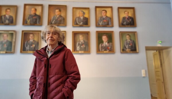 Äldre kvinna framför vägg med porträtt av officerare.