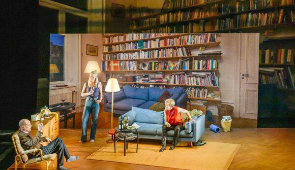 en man och två kvinnor på en teaterscen som föreställer ett vardagsrum med en enorm bokhylla