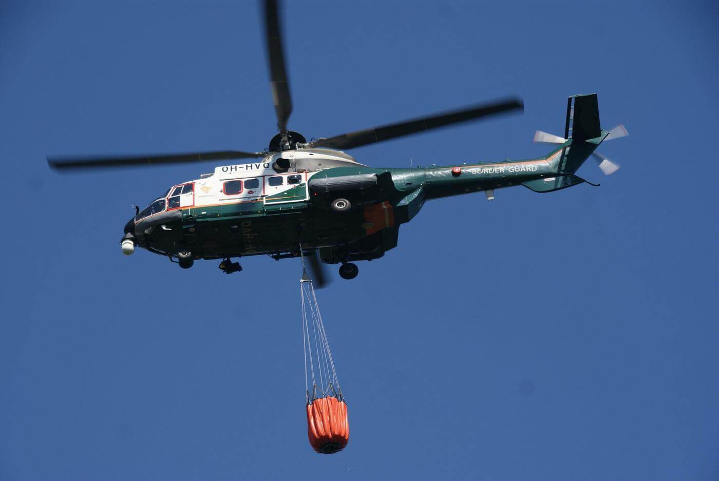 Helikopter i luften med orange påse hängande under sig
