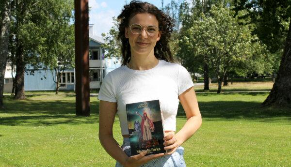 En ung kvinna står utomhus i en grön park. Hon tittar in i kameran och ler i handen håller hon upp en bok.