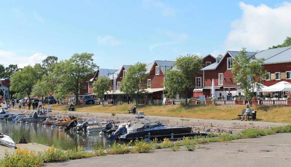 Vatten och några båtar som står vid bryggan i Östra hamnen i Hangö. röda trähus syns längs med gatan som går längs med vattnet. Många människor sitter på husens terrasser.