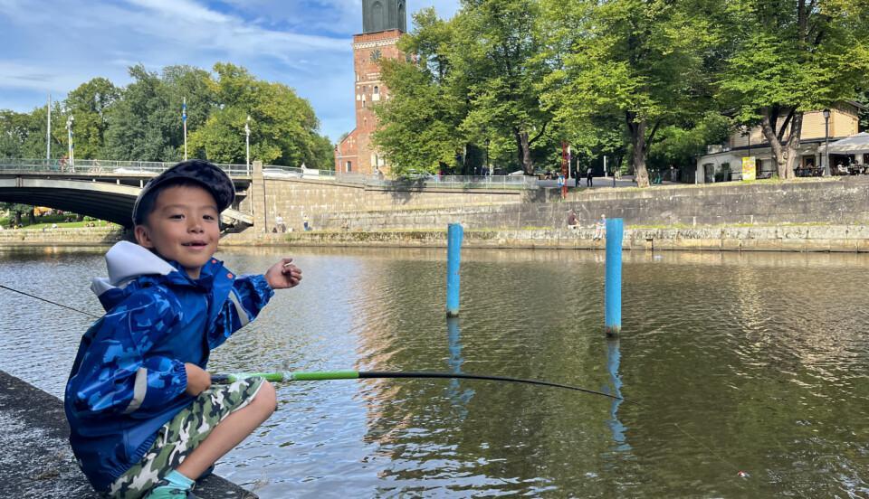 Ung kille med fiskespö sitter på huk vid en å.