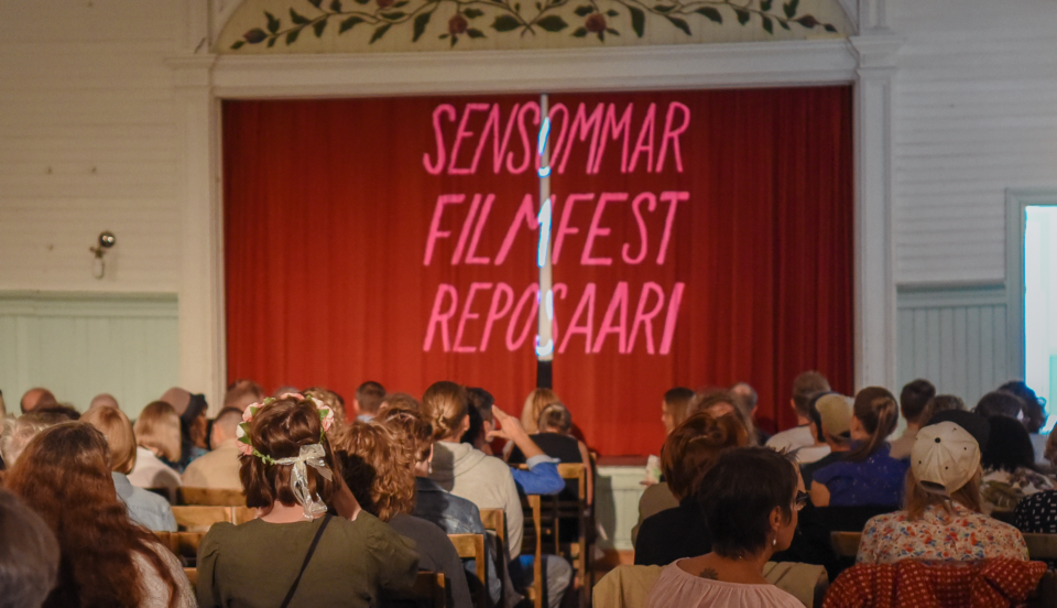 Gammaldags festsal med publik som sitter framför en scen med röd ridå fördragen. På ridån projiceras texten "Sensommar filmfest Reposaari".